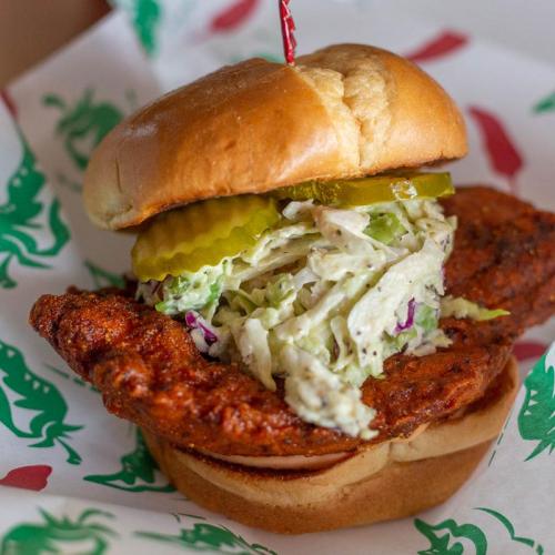 A crispy fried chicken sandwich with pickles and coleslaw on a soft bun, served on a decorative paper.