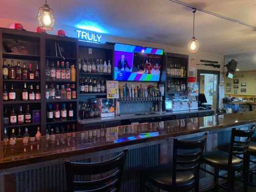 A bar with a wooden counter, shelves of liquor, and a TV displaying a sports event. Neon sign reads "TRULY."