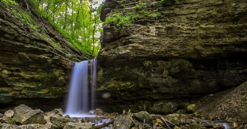 A serene waterfall cascades over rocky cliffs, surrounded by lush green trees in a tranquil forest setting.