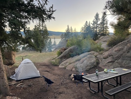 A serene campsite with a tent, picnic table, and a small fire, surrounded by trees and a lake at sunset.