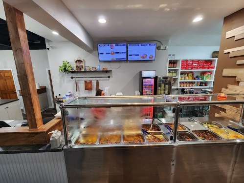 A view of a restaurant counter with food trays, menus on screens, and a clean, modern interior.