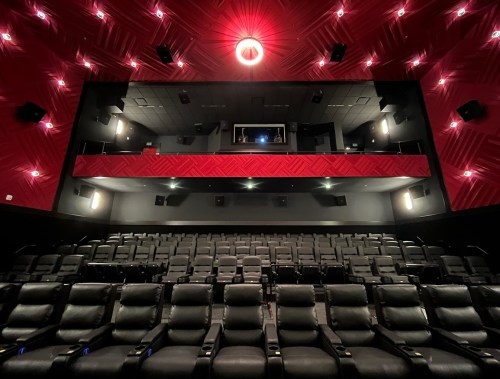 Interior of a modern movie theater featuring black reclining seats and a red accent wall, with a projection booth above.