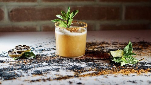 A cocktail garnished with mint sits on a table sprinkled with spices and dried lime slices.