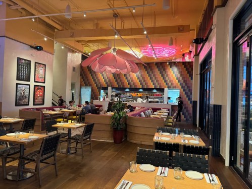 Modern restaurant interior with colorful decor, a large flower light fixture, and a vibrant open kitchen.