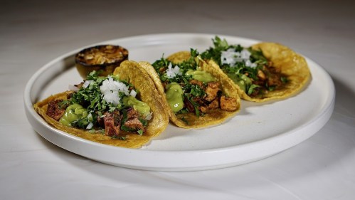 A plate of four tacos filled with meat, onions, cilantro, and guacamole, accompanied by a grilled lime.
