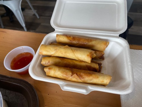 A takeout container holding five crispy spring rolls with a small cup of dipping sauce on the side.
