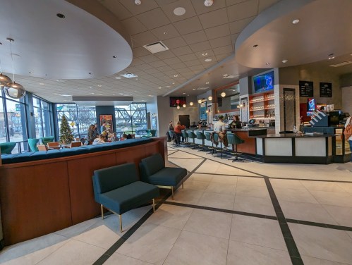 Modern café interior with seating areas, a bar, and large windows. People are enjoying their time inside.