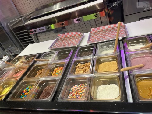 A variety of ice cream toppings in containers, including nuts, sprinkles, and candies, displayed in a dessert shop.