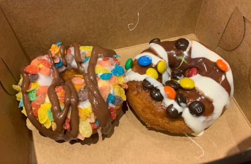 Two colorful donuts in a box: one topped with chocolate and cereal, the other with chocolate and candy-coated chocolates.