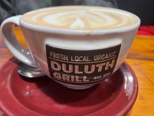 A large cup of latte with intricate foam art, featuring a logo for Duluth Grill on the side.