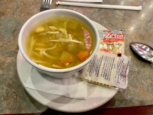 A bowl of chicken noodle soup with vegetables, served with a packet of crackers on a plate.