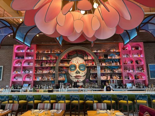 Colorful bar with a large decorative skull mural, shelves of liquor, and vibrant floral ceiling design in a lively restaurant.