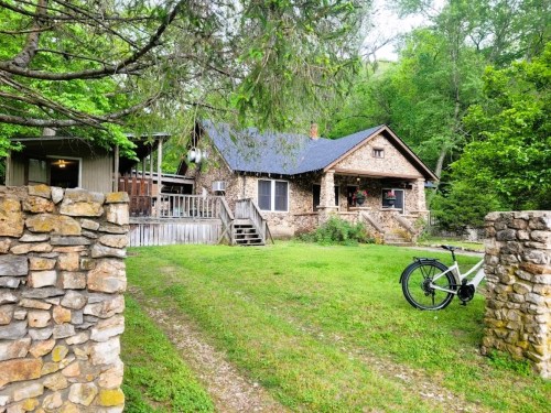 A stone house surrounded by greenery, with a bike parked in front and a gravel path leading to the entrance.