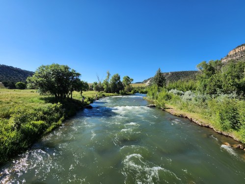 A serene river flows through a lush green landscape under a clear blue sky, surrounded by trees and hills.