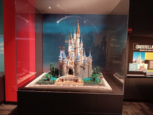 A detailed LEGO model of Cinderella's Castle displayed in a glass case, with a starry backdrop and greenery.