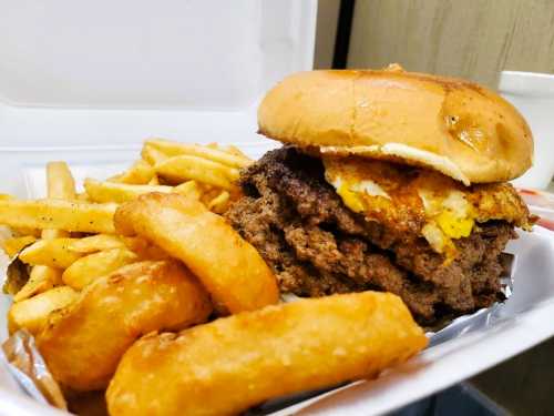 A hearty burger with a fried egg on top, served with crispy fries and potato wedges in a takeout container.