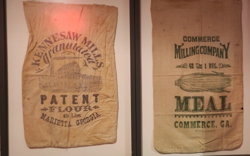 Two vintage flour and meal sacks displayed on a wall, featuring faded text and illustrations from Kennesaw and Commerce, Georgia.