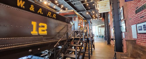 A vintage train car displayed indoors, with stairs leading up to it and industrial decor in the background.
