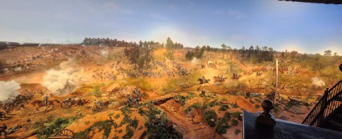 A panoramic view of a historical battle scene with soldiers, horses, and smoke amidst a rugged landscape.