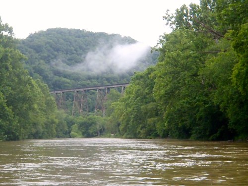 A river flows through lush greenery, with a misty mountain and an old railway bridge in the background.