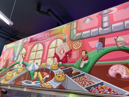 Colorful mural of a candy factory with whimsical characters, donuts, and sweets in a vibrant, playful setting.