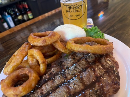A plate with a grilled steak, onion rings, a bun, and a beer from The Fort Bar & Grill.