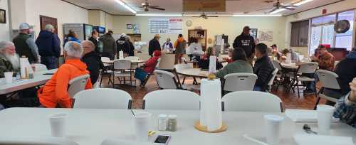 A busy dining area with people seated at tables, some in line, and a counter with food service in the background.