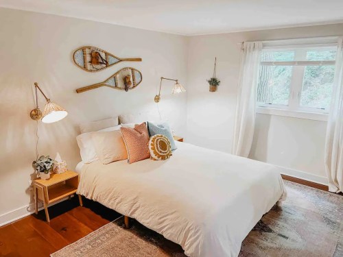 Cozy bedroom with a double bed, decorative pillows, wall sconces, and vintage snowshoes on the wall. Natural light from window.