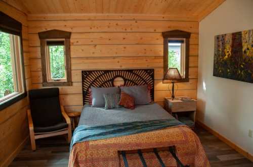 Cozy bedroom with wooden walls, a bed with colorful bedding, a chair, and windows letting in natural light.