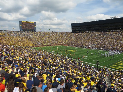A packed stadium filled with fans in maize and blue, watching a football game under a cloudy sky.