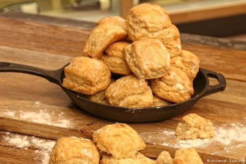 A pile of freshly baked biscuits in a cast iron skillet, with some crumbs and flour scattered on a wooden surface.