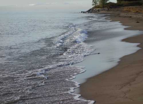 Gentle waves lapping at a sandy beach, with calm water and a distant shoreline under a soft sky.