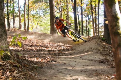 A mountain biker leans into a turn on a dirt trail surrounded by autumn trees. Dust kicks up behind them.