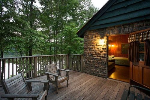 Cozy cabin with a wooden deck overlooking a serene lake, surrounded by lush trees. Warm light glowing from inside.