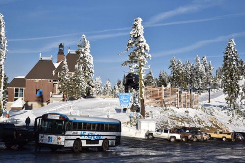 A snowy landscape featuring a lodge, trees, and parked vehicles, with a blue sky in the background.