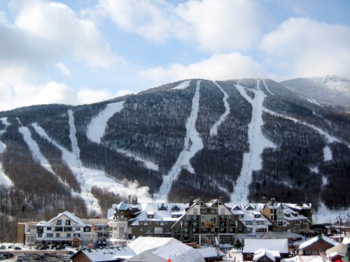 A snowy mountain with ski trails and a resort at its base under a partly cloudy sky.