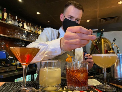A bartender in a mask prepares cocktails at a bar, with four drinks displayed in front of him.