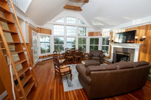 Bright, airy living room with large windows, wooden accents, a fireplace, and a dining area overlooking a lake.