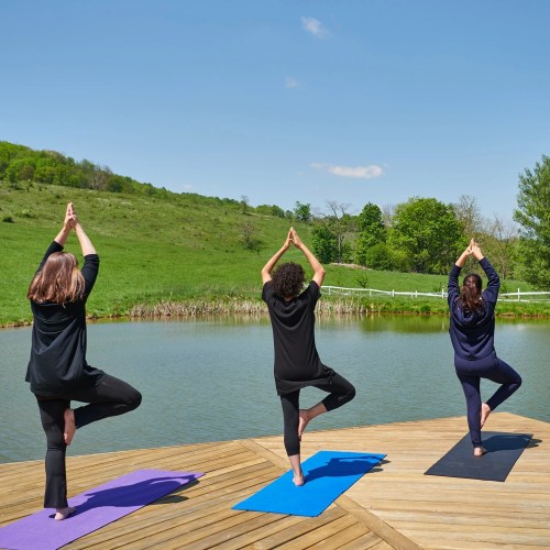 Three people practicing yoga on a dock by a serene pond, surrounded by green hills and a clear blue sky.