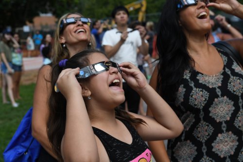 A group of people, including a girl, wearing eclipse glasses and looking up, excited during an outdoor event.