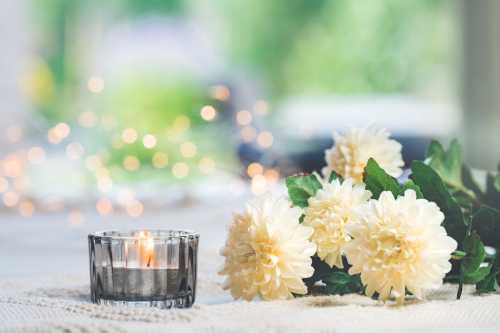 A lit candle beside soft white flowers on a textured surface, with blurred lights in the background.