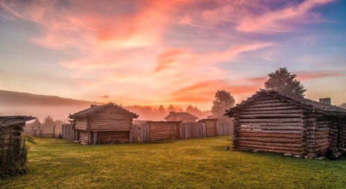 A serene landscape featuring rustic log cabins at sunrise, surrounded by mist and lush green grass.