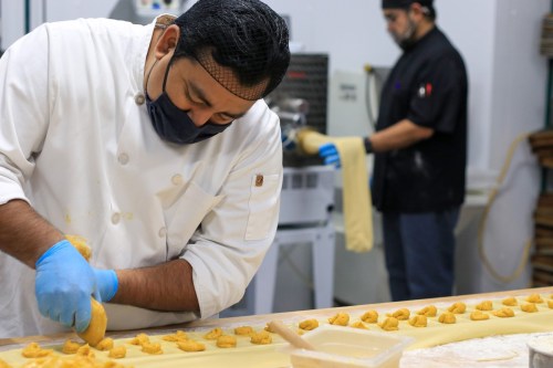 A chef in a white coat pipes dough onto a countertop, while another chef works in the background.