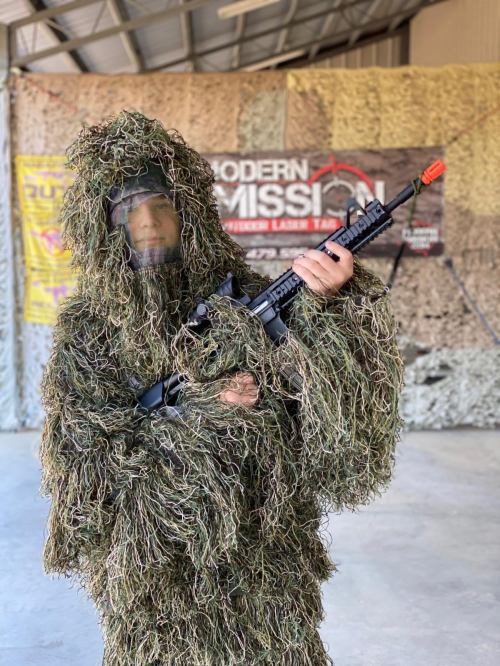 A person in a camouflage ghillie suit holds a rifle, standing in a military-style indoor setting.