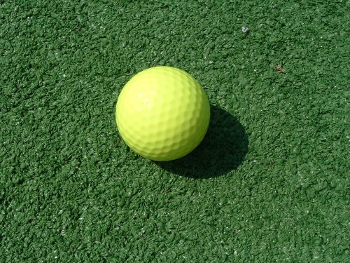 A bright yellow golf ball resting on a green artificial turf surface.