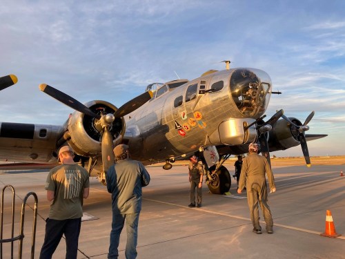 A vintage bomber aircraft on a runway, with crew members inspecting it under a clear sky at sunset.
