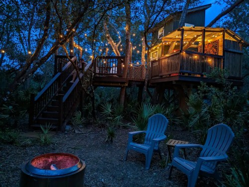A cozy treehouse illuminated by string lights, surrounded by trees, with a fire pit and chairs in the foreground.