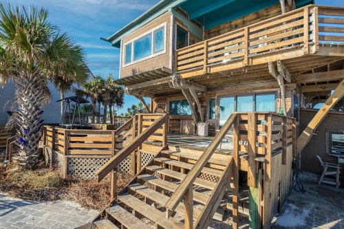 A wooden beach house with a deck and stairs, surrounded by palm trees and a clear blue sky.