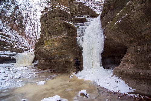 A person stands near a frozen waterfall in a rocky, snow-covered canyon surrounded by trees.