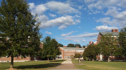 A sunny campus scene with brick buildings, green trees, and a blue sky filled with fluffy clouds.